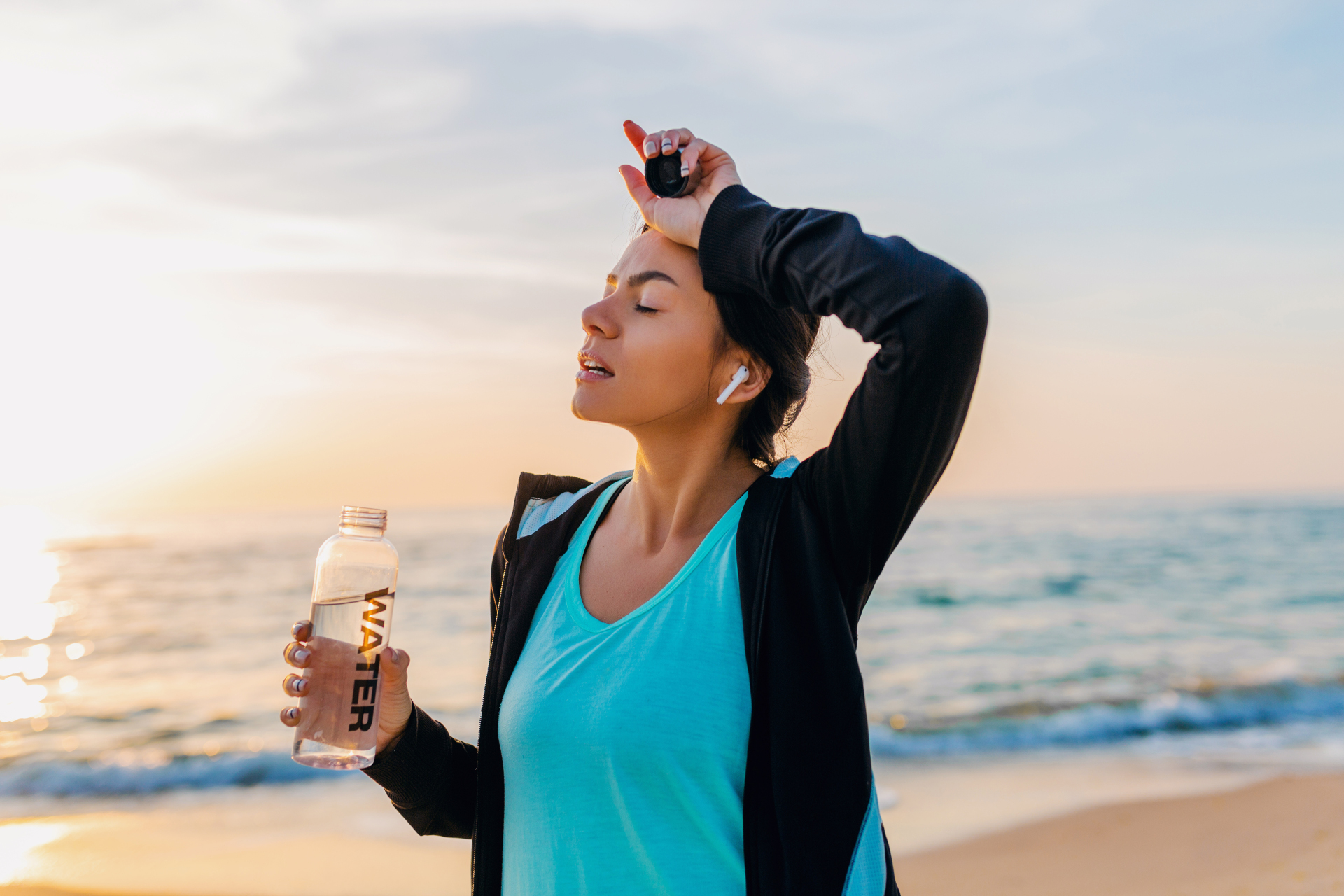 attractive slim woman doing sport exercises on morning sunrise beach in sports wear, thirsty drinking water in bottle, healthy lifestyle, listening to music on wireless earphones, smiling happy